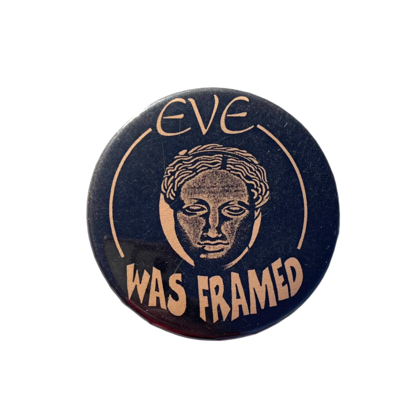 A round black badge with pale orange text , featuring the phrase 'EVE WAS FRAMED' and an illustration of a female head.