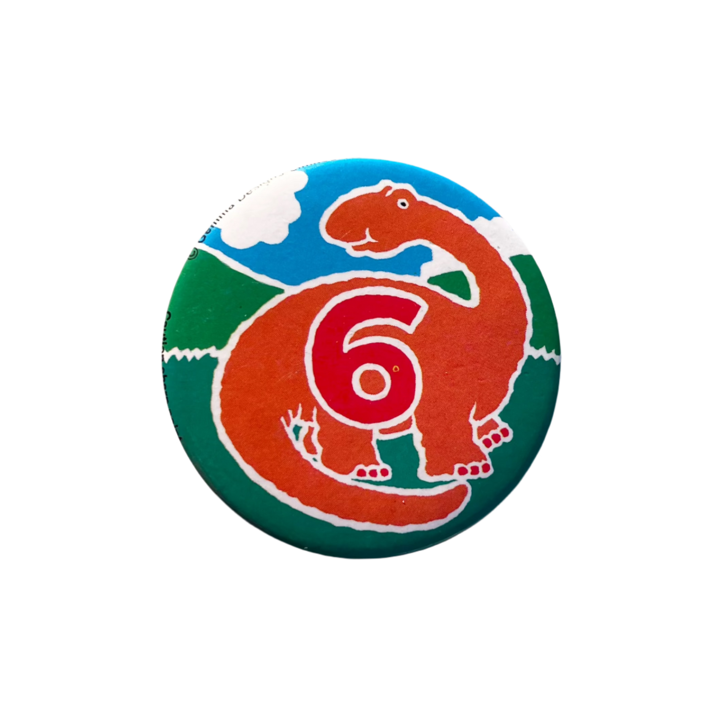 A round birthday badge with green grass and a blue cloudy sky featuring an illustration of an orange dinosaur and the number 6 in red.
